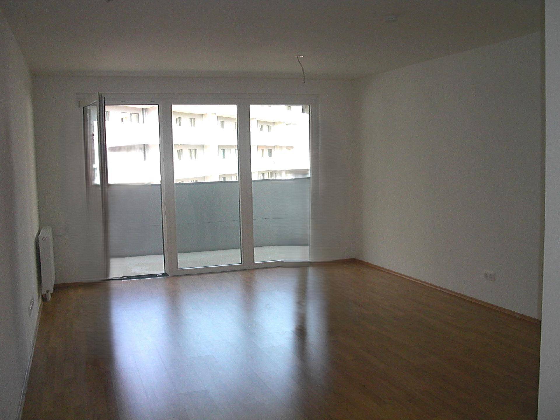 Leeres Wohnzimmer mit hellem Parkettboden und Zugang zu einem Balkon.