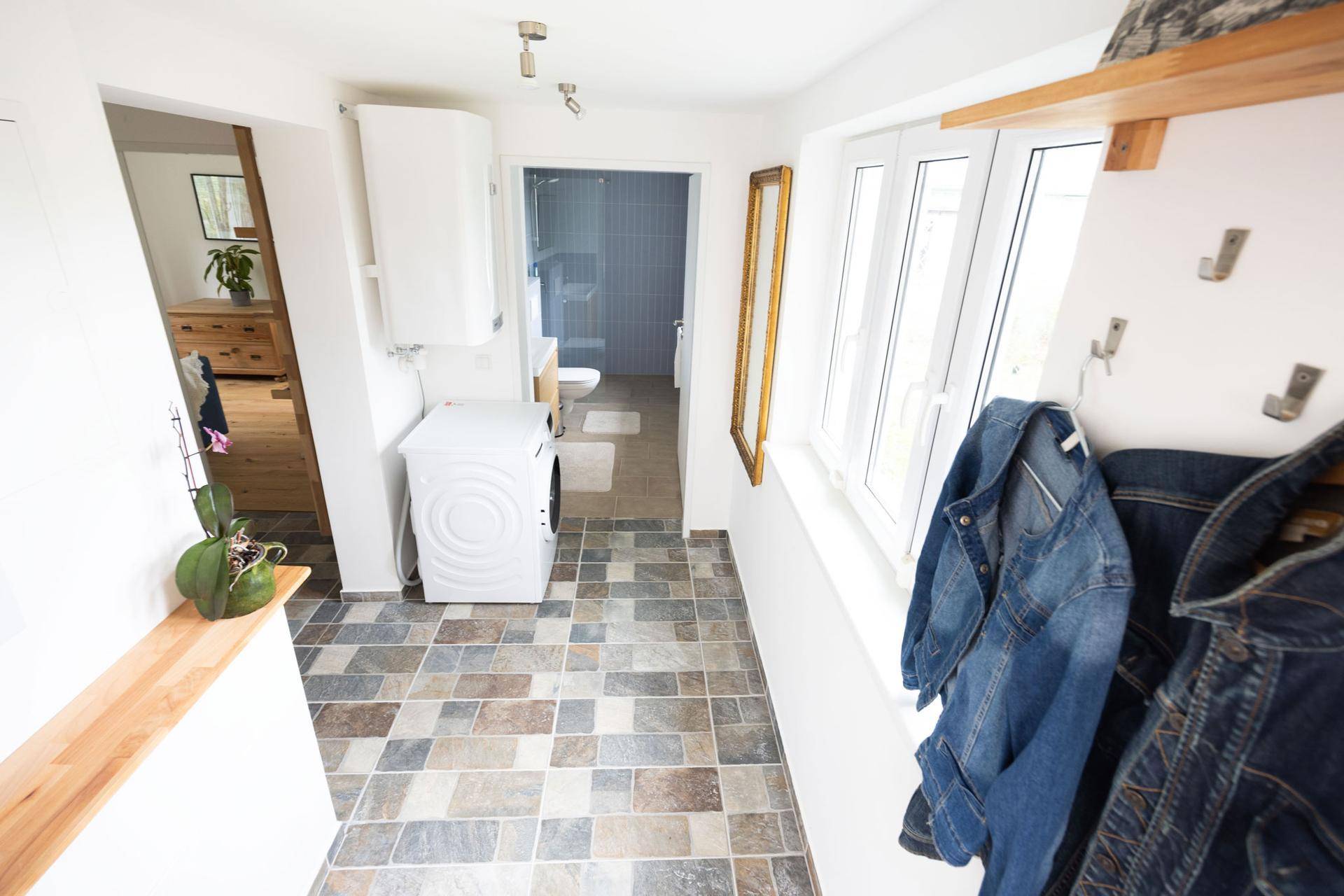 Heller Flur mit Waschmaschine, Warmwasserboiler und Blick in ein gefliestes Badezimmer.