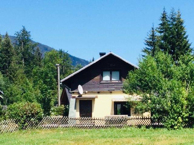 Freistehendes Haus in ländlicher Umgebung mit Wald und Bergen im Hintergrund unter blauem Himmel.