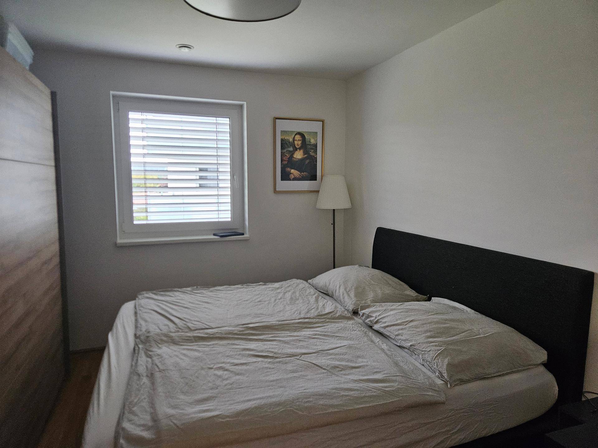 Schlafzimmer mit Doppelbett, Kleiderschrank und Fenster mit Jalousien.