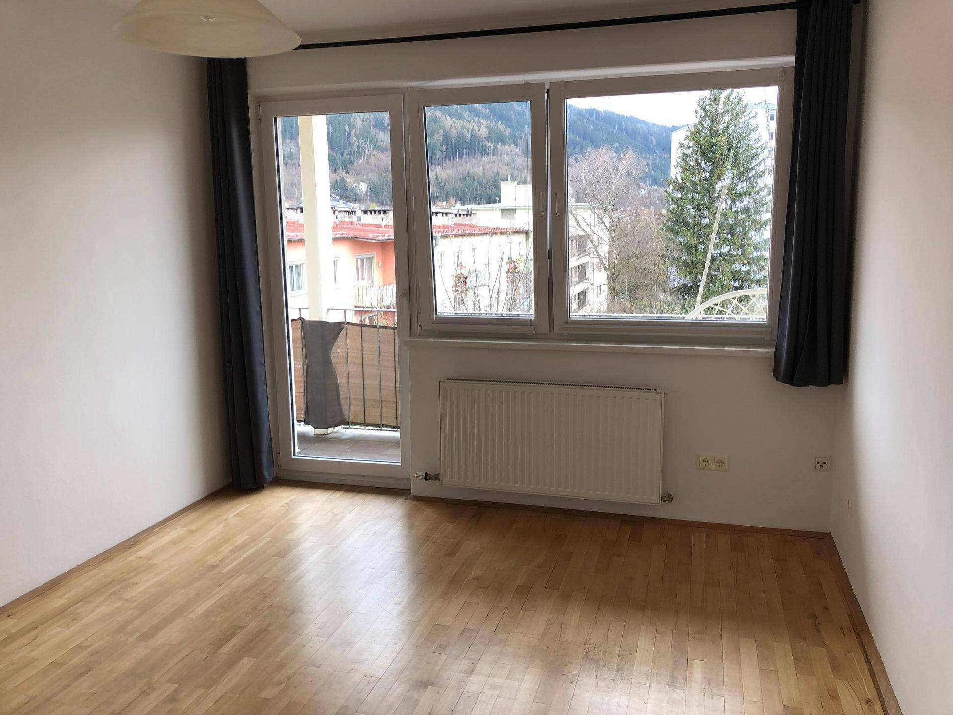 Leeres Schlafzimmer mit Holzboden, großem Fenster und Zugang zu einem kleinen Balkon mit Bergblick.