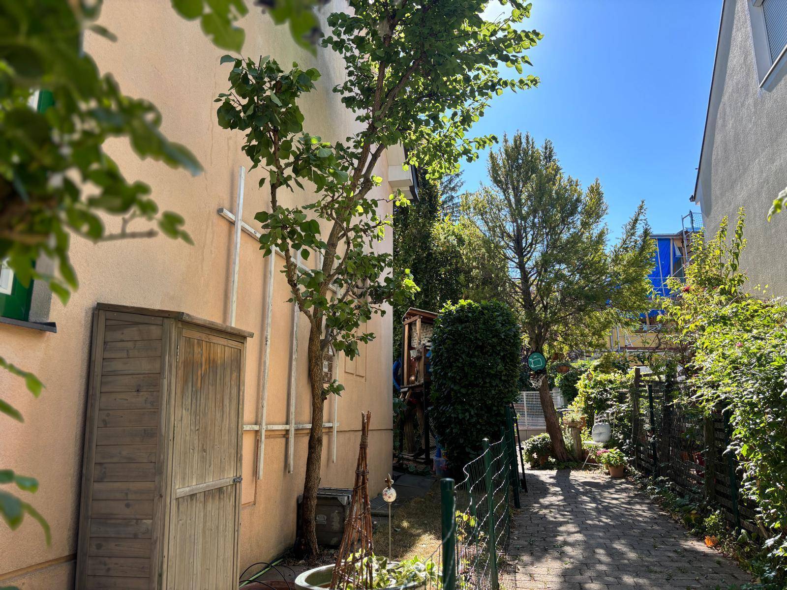 Sonnige Seitenansicht des Hauses mit einem gepflasterten Weg, Bäumen und üppigem Grün im Garten.