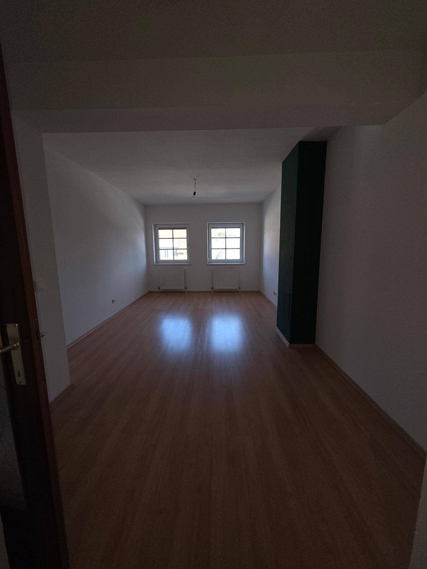 Geräumiges Wohnzimmer mit Laminatboden und zwei Fenstern, die viel Tageslicht hereinlassen.