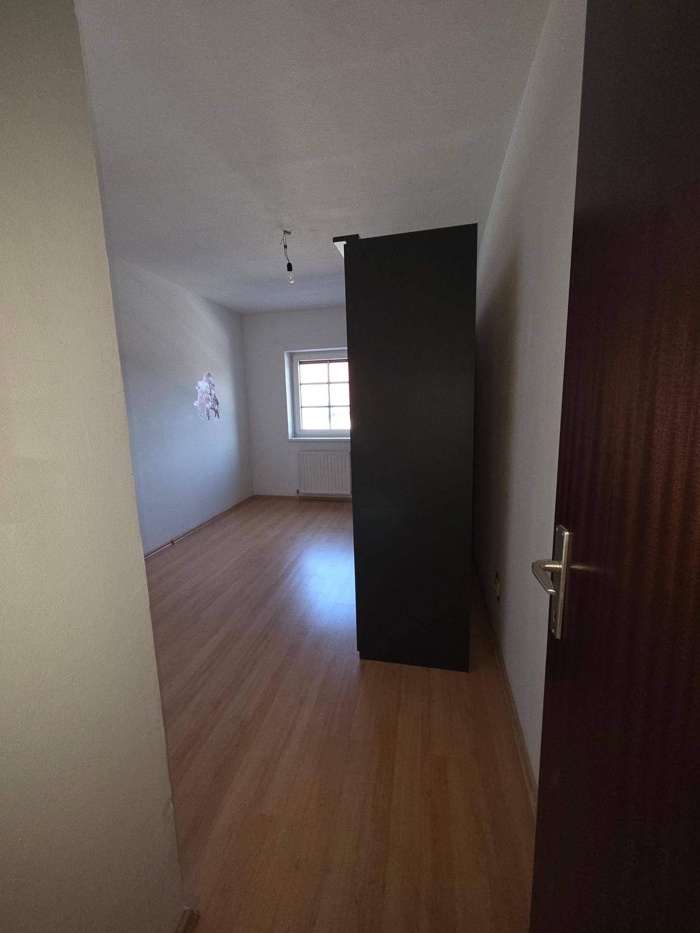 Helles Zimmer mit Laminatboden und einem großen dunklen Schrank, ideal als Schlaf- oder Arbeitszimmer.