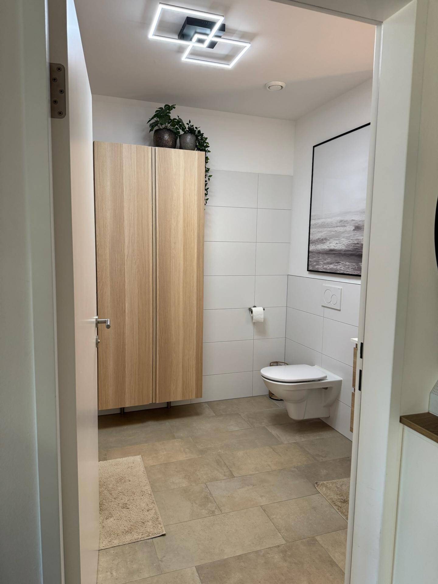 Modernes Badezimmer mit Wand-WC, hohem Holzschrank und hellen Wand- und Bodenfliesen.