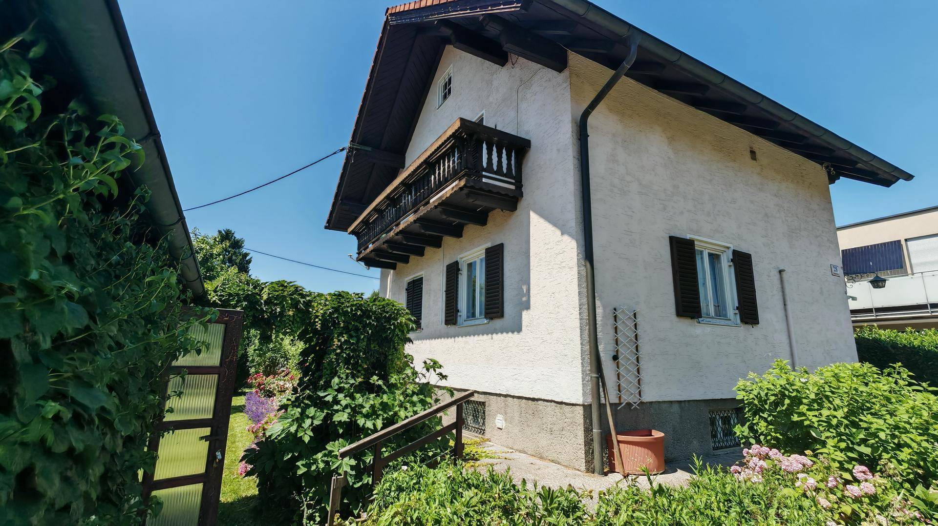Seitenansicht des Hauses mit traditionellem Holzbalkon, Satteldach und grüner Umgebung unter blauem Himmel.