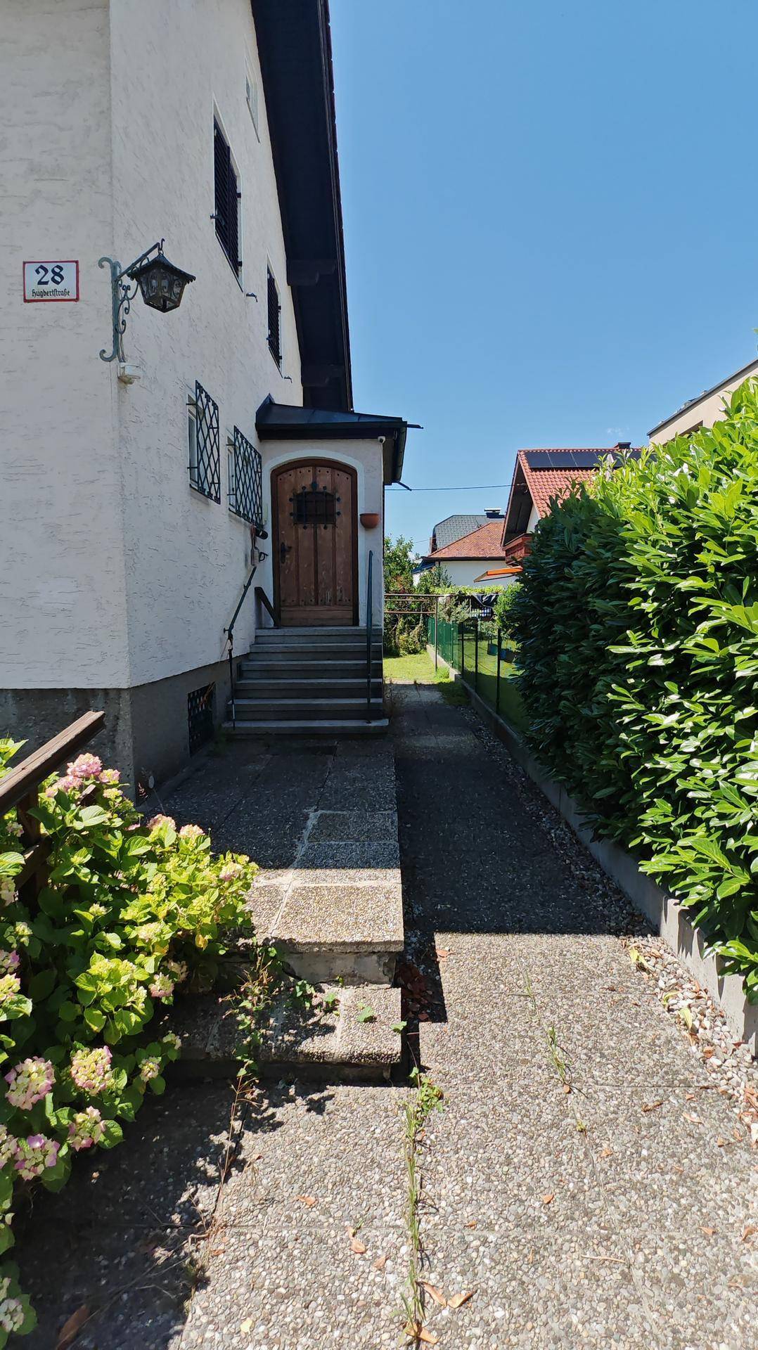 Eingangsbereich des Hauses mit Steintreppe, Holztür und gepflastertem Weg, umgeben von Hecken.