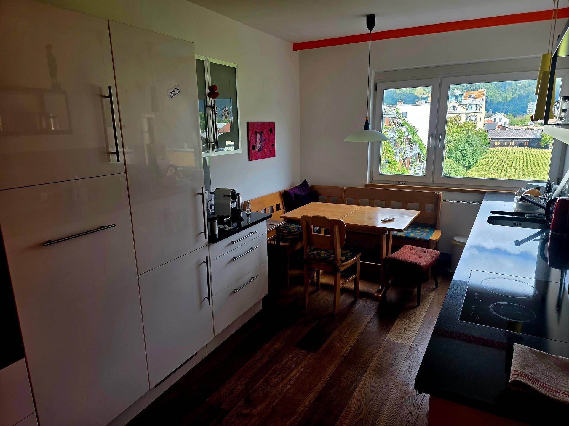 Moderne Küche mit weißen Schränken, dunkler Arbeitsplatte und gemütlicher Essecke mit Blick auf Weinberge.