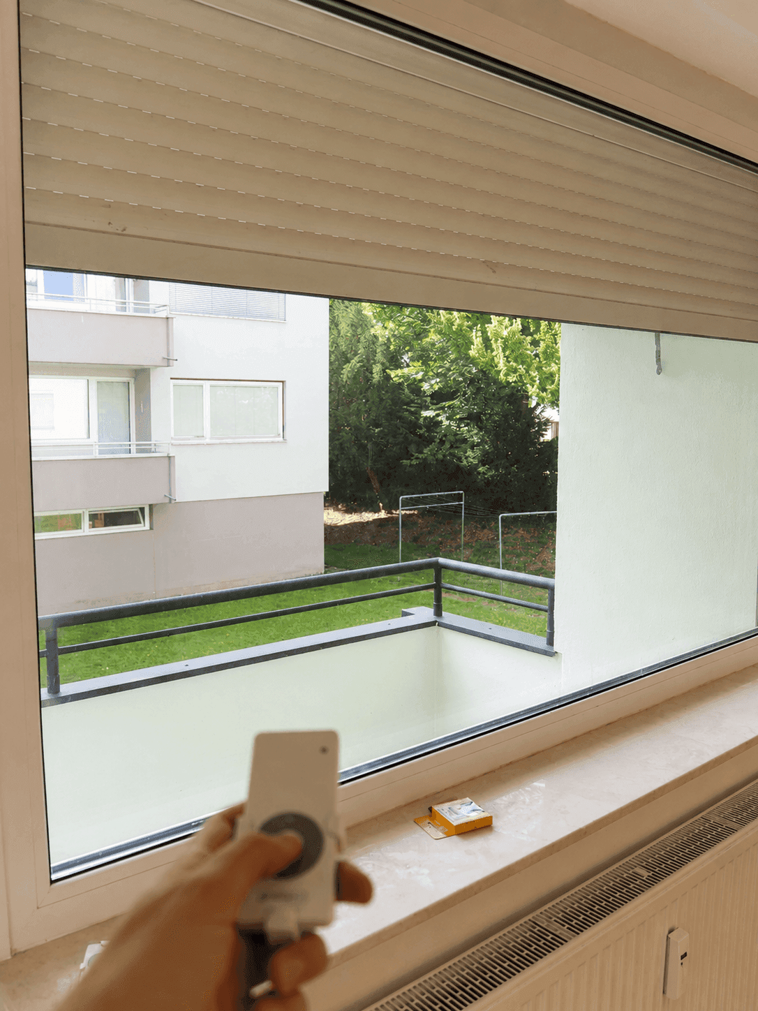 Fenster mit teilweise geöffnetem Rollladen und Blick auf die grüne Umgebung, gesteuert per Fernbedienung.