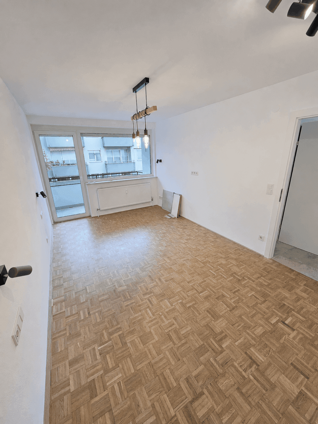 Heller Wohnraum mit Fischgrätparkett und großem Fenster, das Zugang zu einem Balkon und viel Tageslicht bietet.
