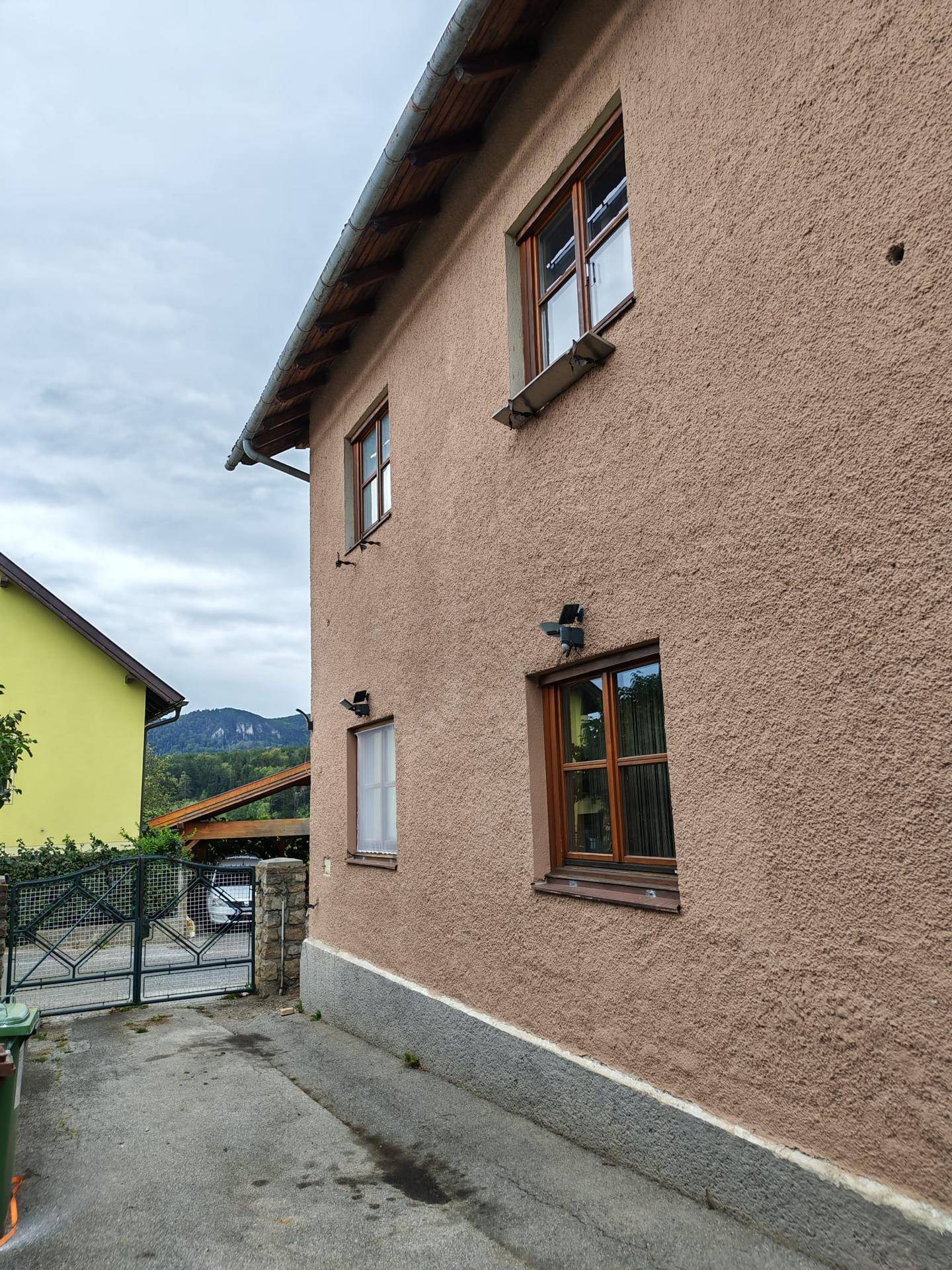 Seitenansicht eines Wohnhauses mit beigefarbener Fassade und braunen Holzfenstern, daneben eine Einfahrt.