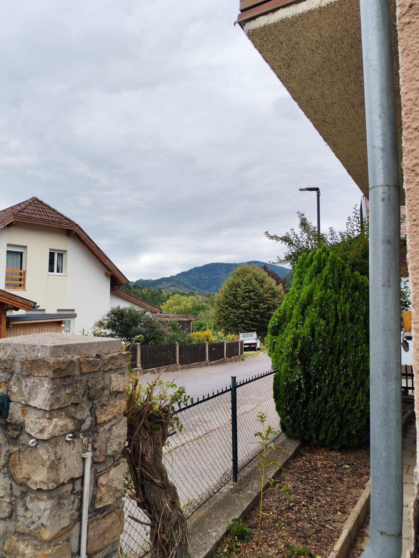 Blick von der Grundstücksgrenze auf eine ruhige Wohnstraße und die umliegende grüne Hügellandschaft.