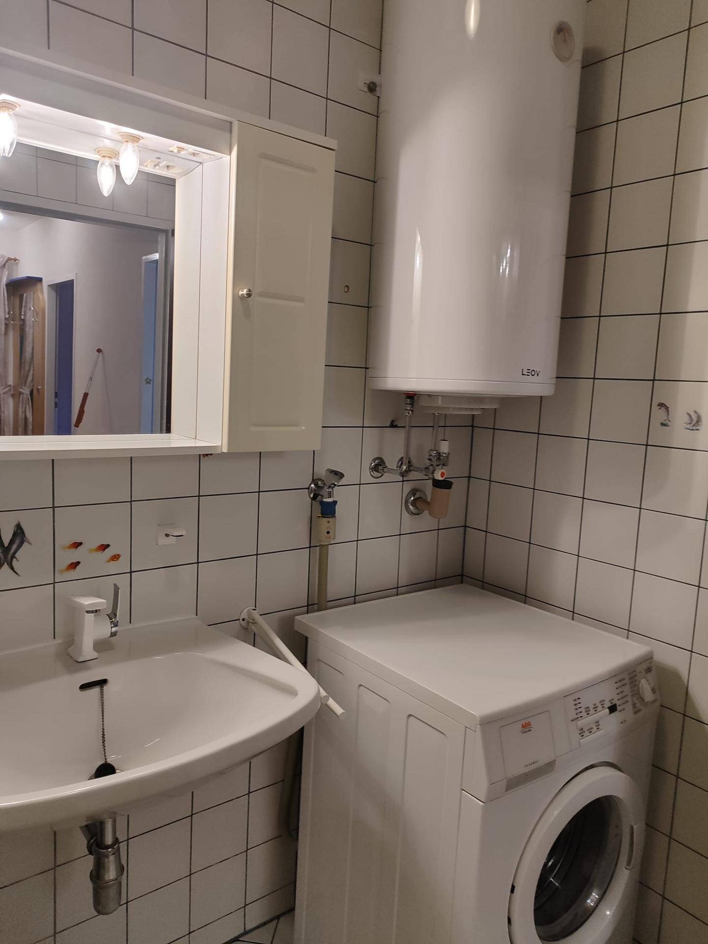 Badezimmer mit Waschmaschine, Waschbecken, Boiler und weiß gefliesten Wänden.