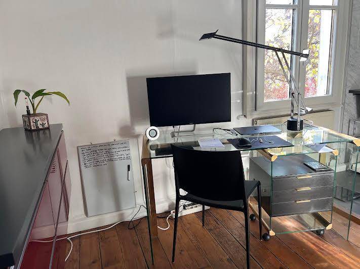 Helles Arbeitszimmer mit Holzboden, modernem Glas-Schreibtisch und großem Fenster.
