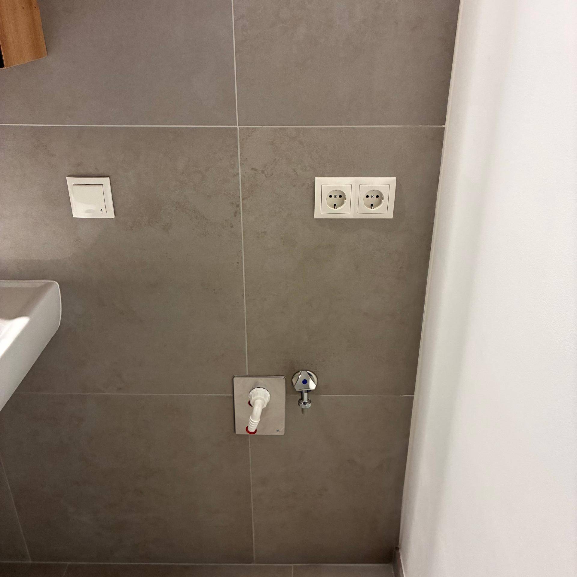 Detailansicht der Badezimmerwand mit grauen Fliesen und Anschlüssen für eine Waschmaschine.