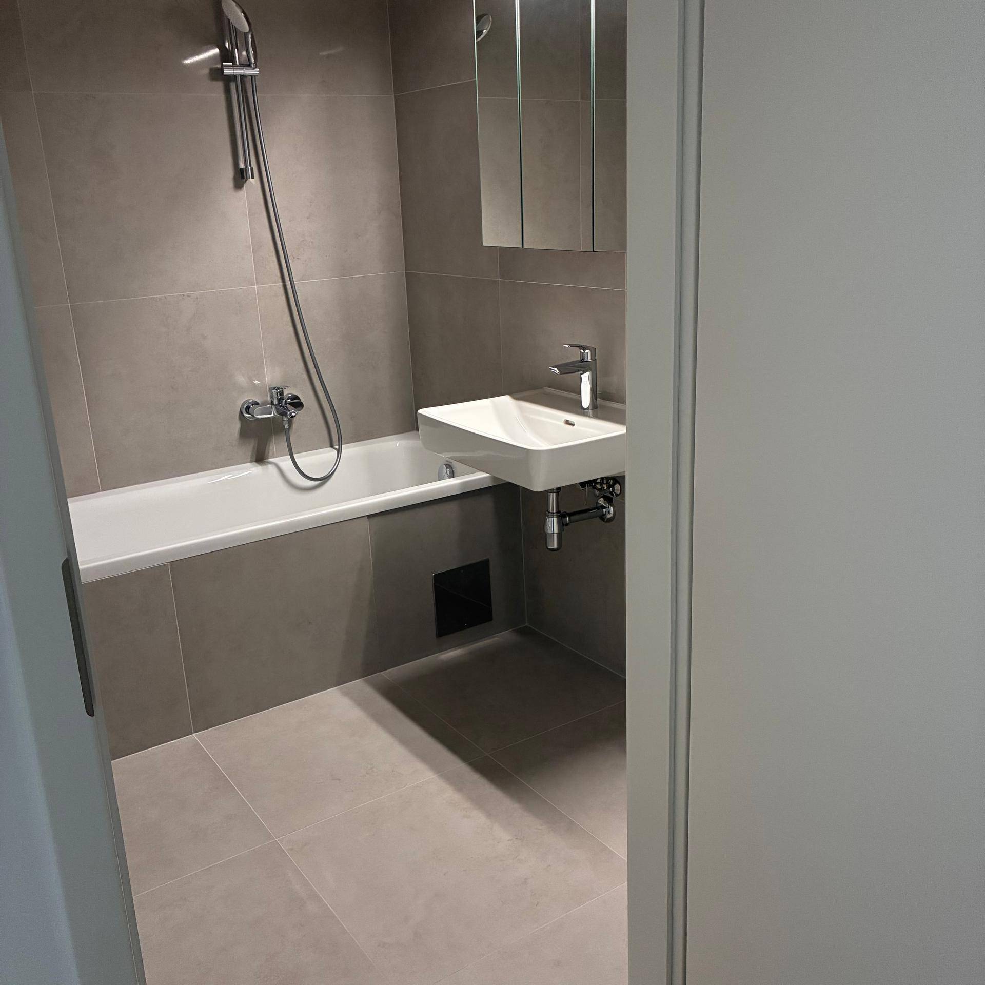 Modernes Badezimmer mit grauen Fliesen, Badewanne mit Duschfunktion und Einzelwaschbecken.