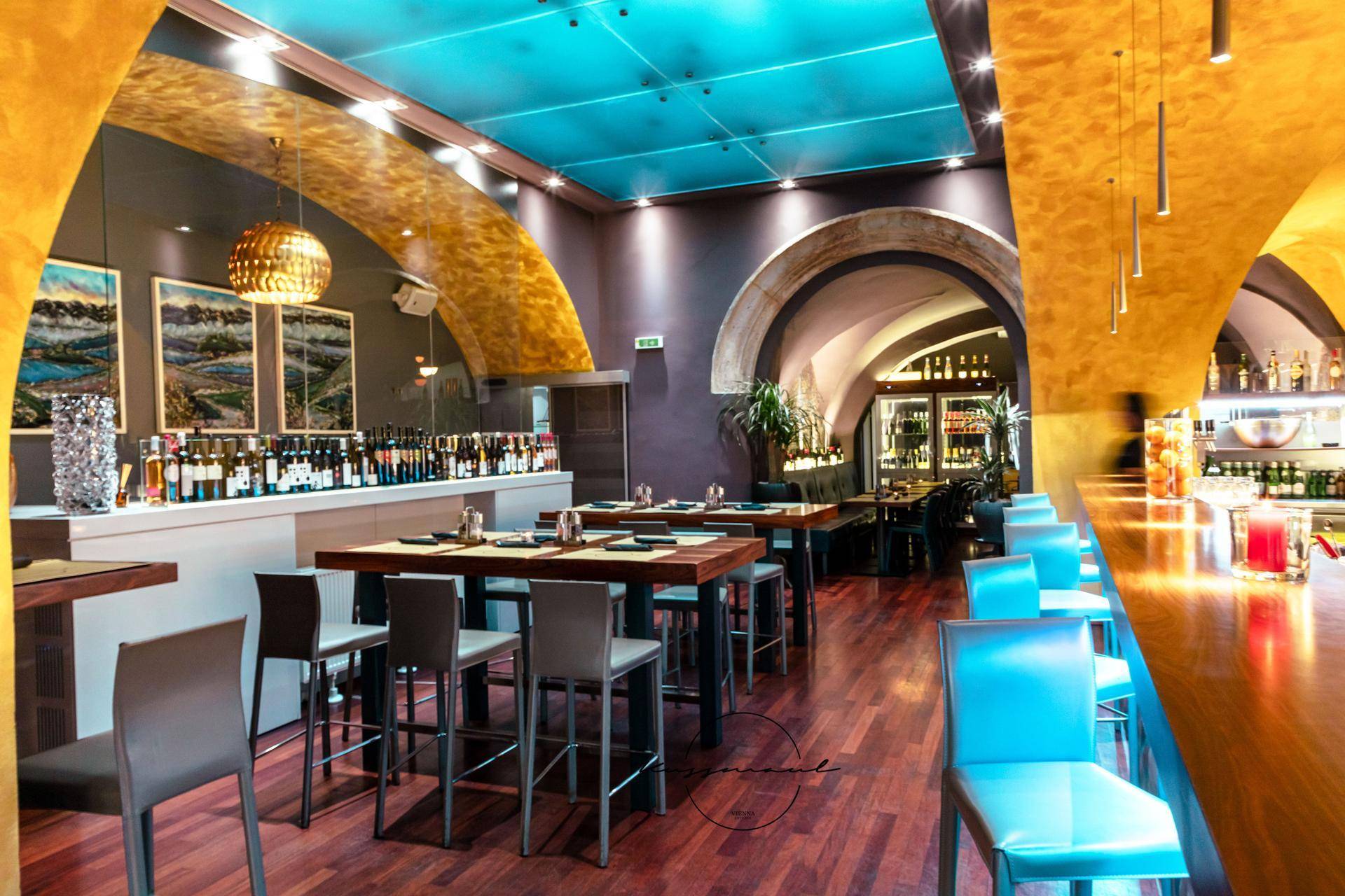 Geräumige Bar mit Holzboden, hohen Tischen und einer Decke mit blauer Akzentbeleuchtung in einem historischen Raum.