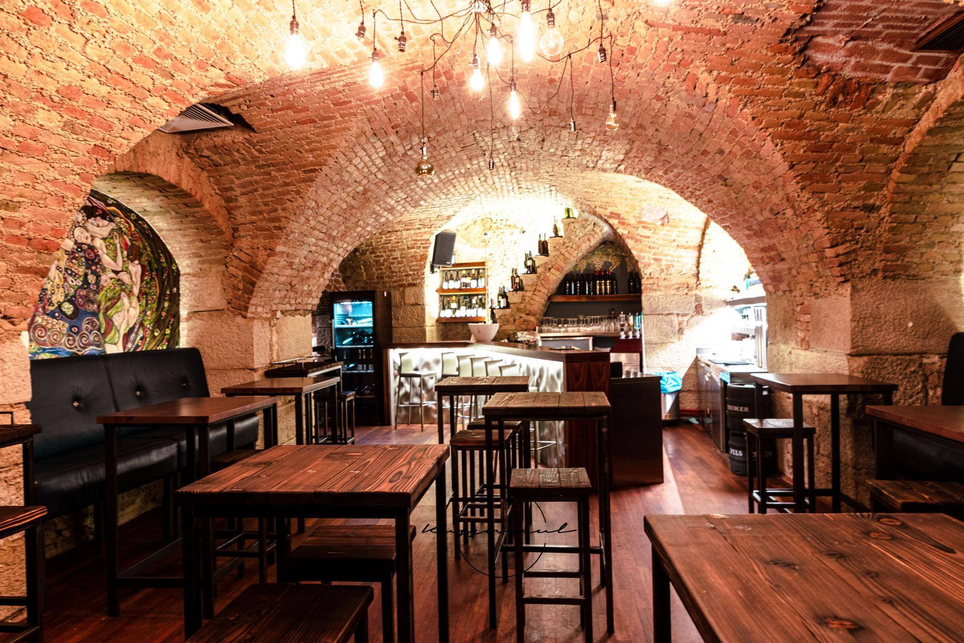 Rustikaler Kellerraum mit Ziegelgewölbe, Holztischen und einer Bar im Hintergrund, ideal für gesellige Abende.