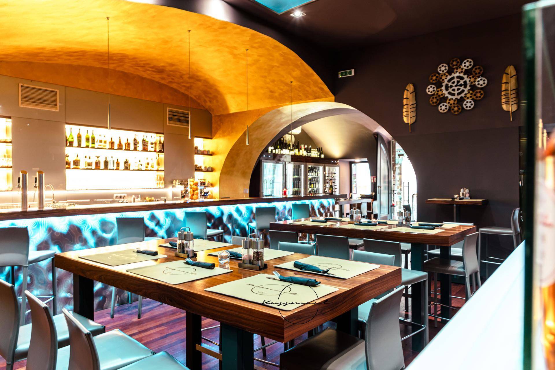 Restaurantbereich mit Holztischen, Barhockern und einer beleuchteten Bar im Hintergrund unter einem Gewölbe.