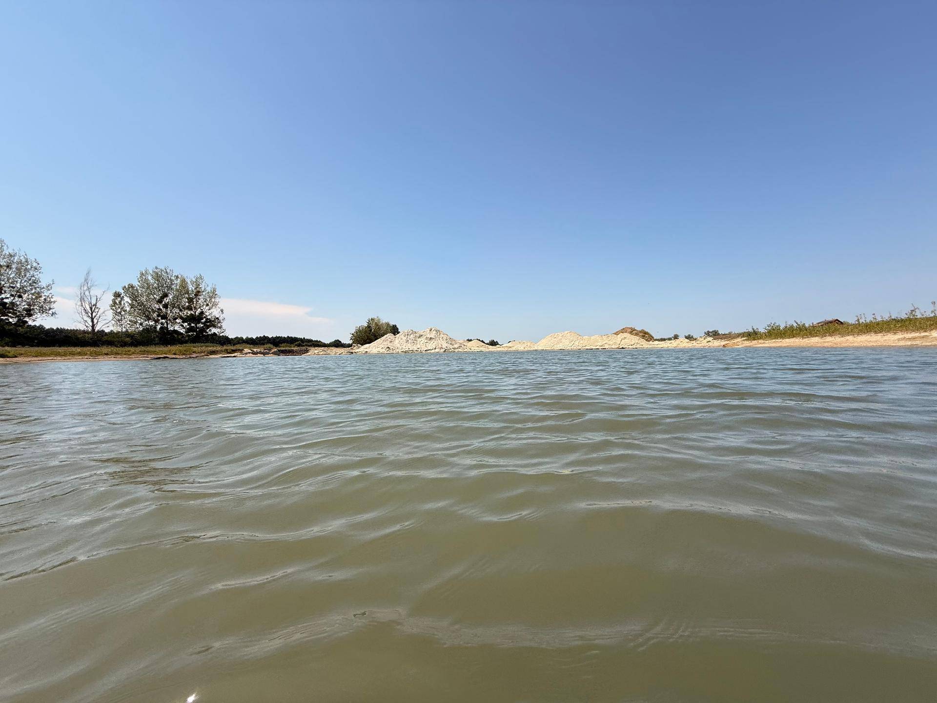 Blick über das Wasser auf sandige Ablagerungen und vereinzelte Bäume am Horizont unter blauem Himmel.