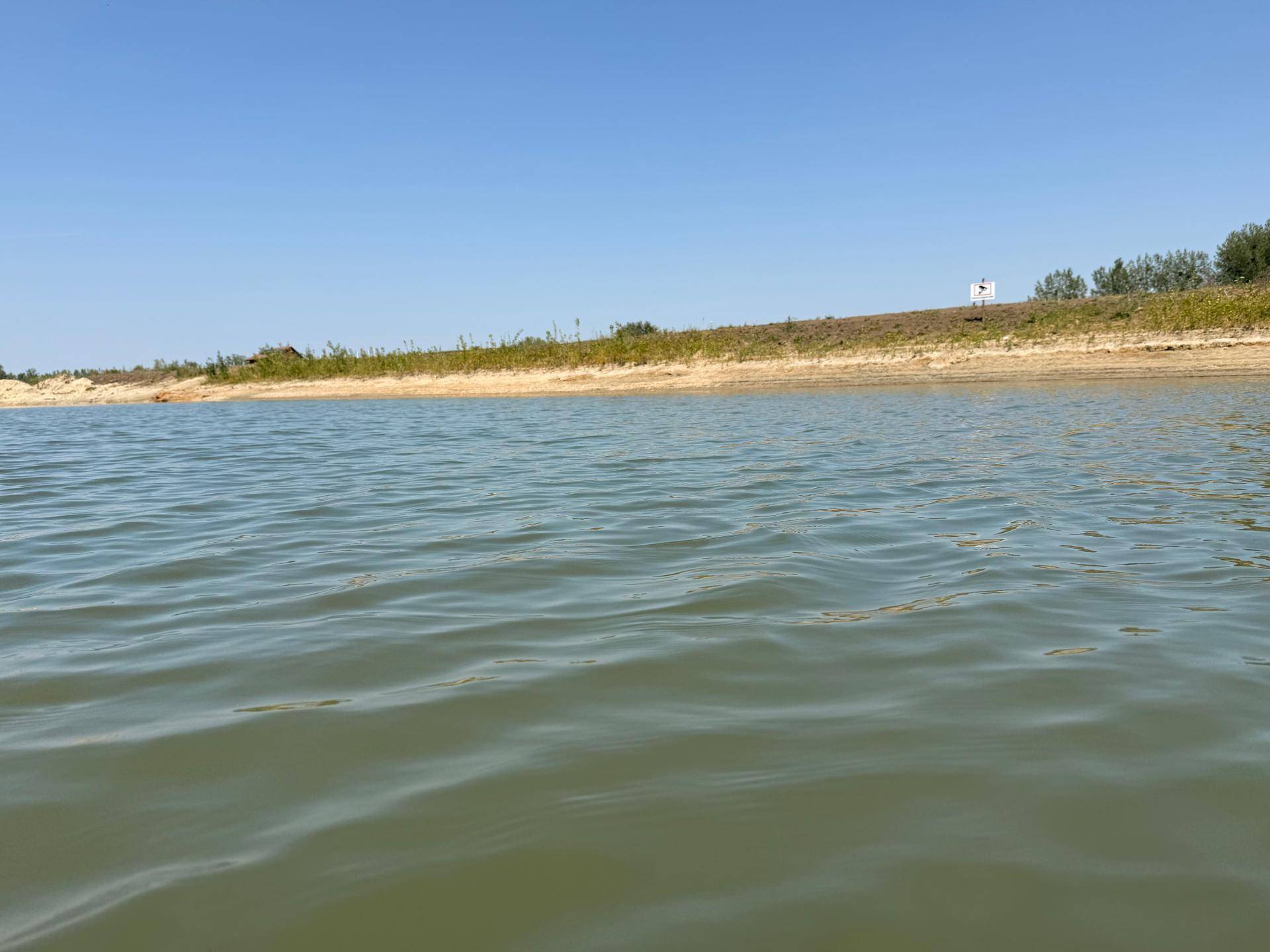 Nahaufnahme der Wasseroberfläche eines Sees mit sandigem Ufer und Vegetation im Hintergrund unter klarem Himmel.