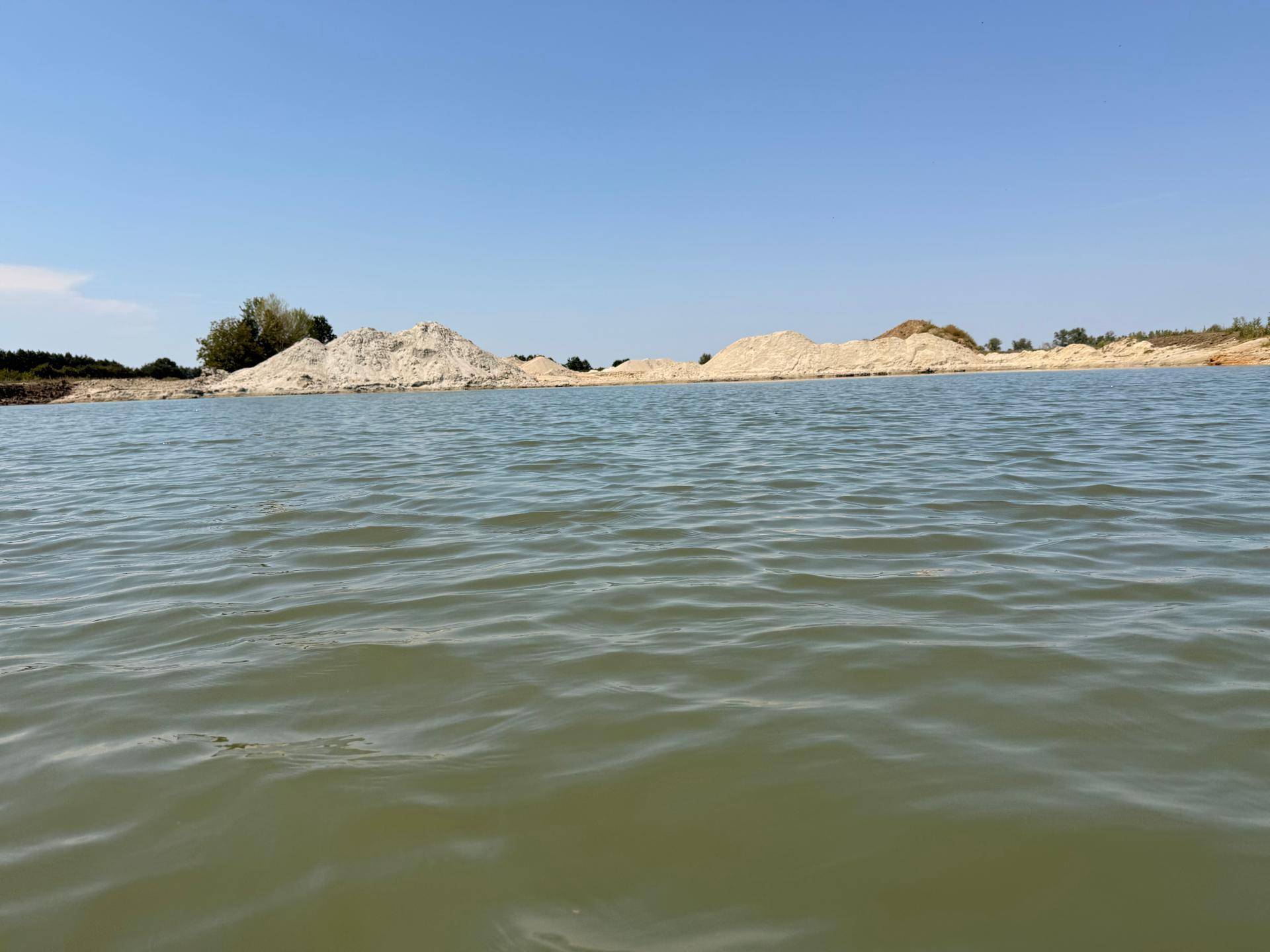 Blick vom Wasser auf sandige Hügel am gegenüberliegenden Ufer unter klarem, blauem Himmel.