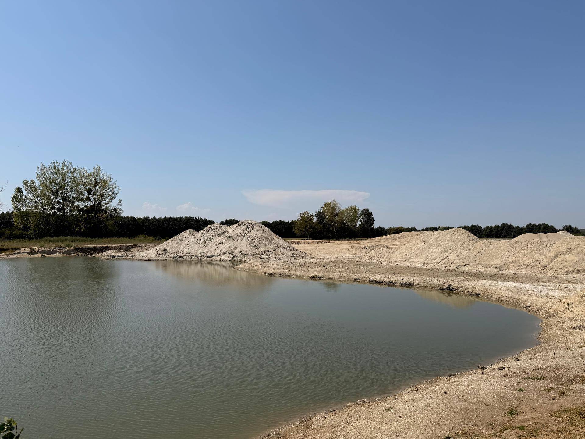Weitläufiger Blick auf einen Teich mit großen Sandhügeln am Ufer und Bäumen im Hintergrund an einem sonnigen Tag.