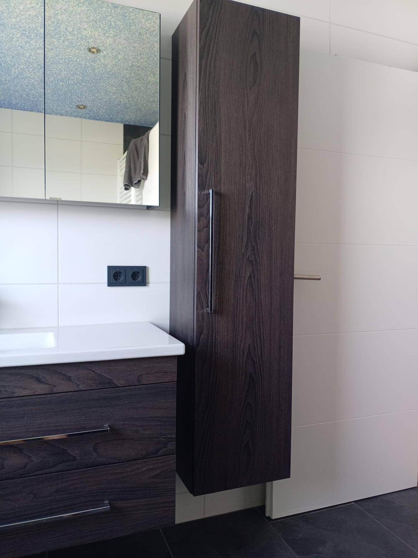 Dunkler Holzschrank im Badezimmer mit Spiegelschrank und weißer Waschtischplatte.