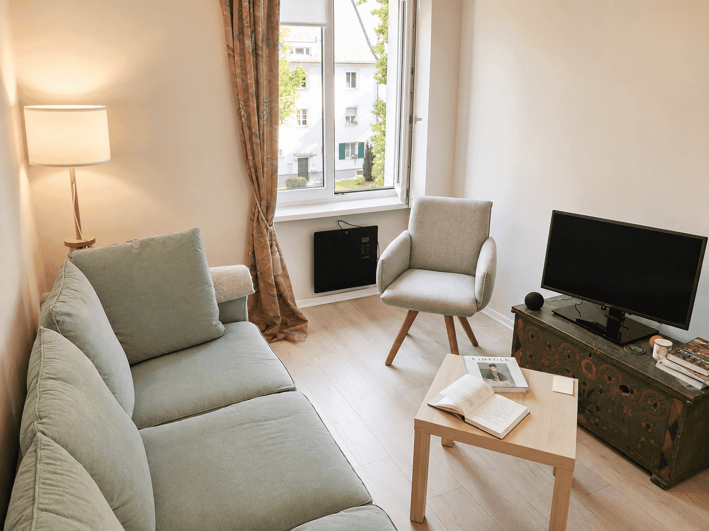 Gemütliches Wohnzimmer mit hellem Sofa, Sessel und einem kleinen Couchtisch vor dem Fenster.