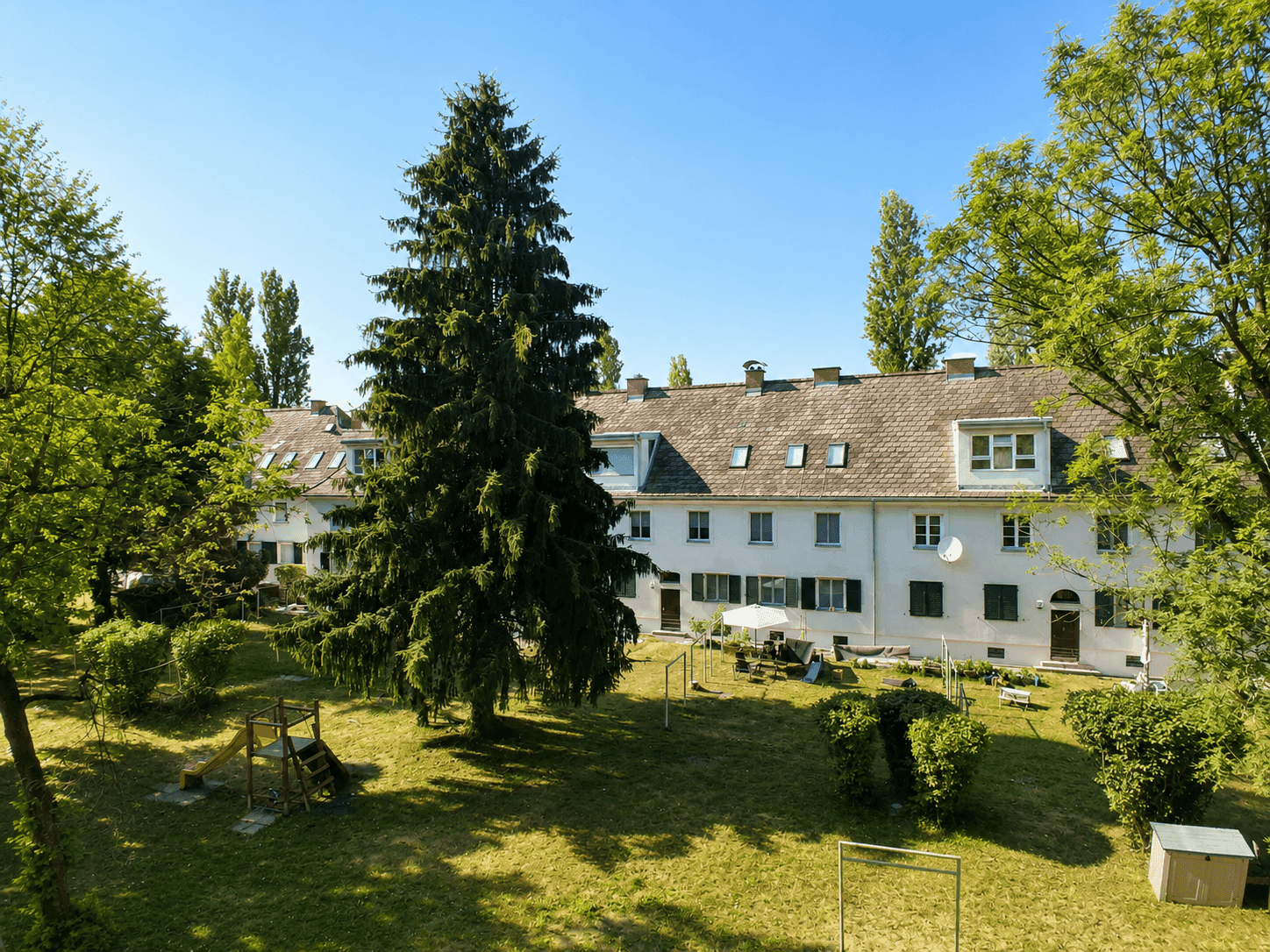 Großer Gemeinschaftsgarten mit Rasenfläche, hohen Bäumen und einem Spielplatz vor den Reihenhäusern.
