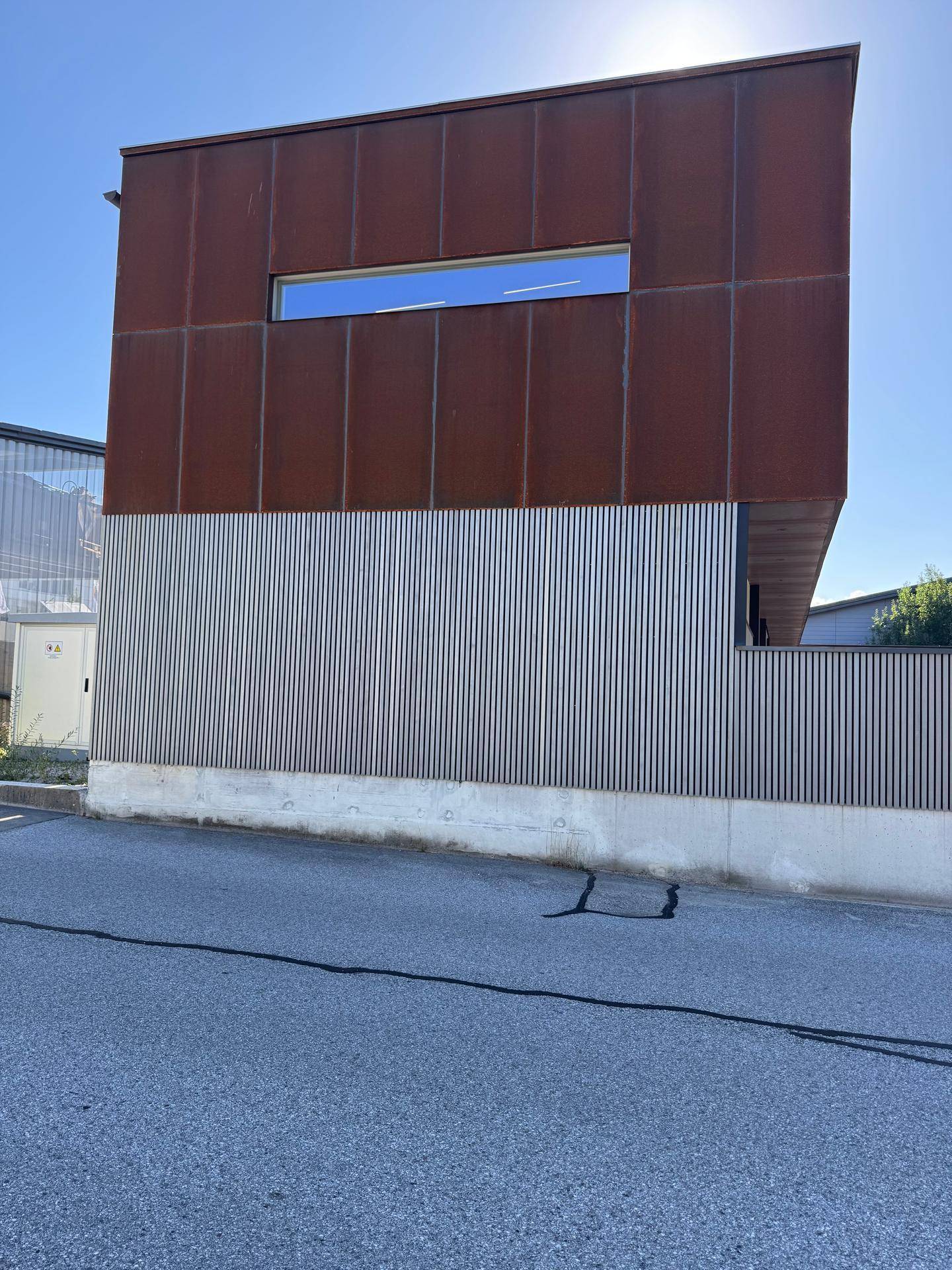 Seitenfassade des Gebäudes mit rostfarbenen Metallplatten und vertikalen Lamellen unter strahlendem Sonnenschein.