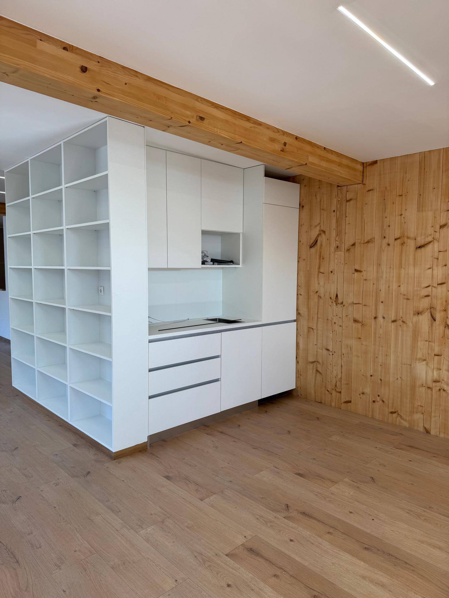 Heller Innenraum mit moderner weißer Küchenzeile, Holzvertäfelung und Holzboden, sowie sichtbarem Deckenbalken.