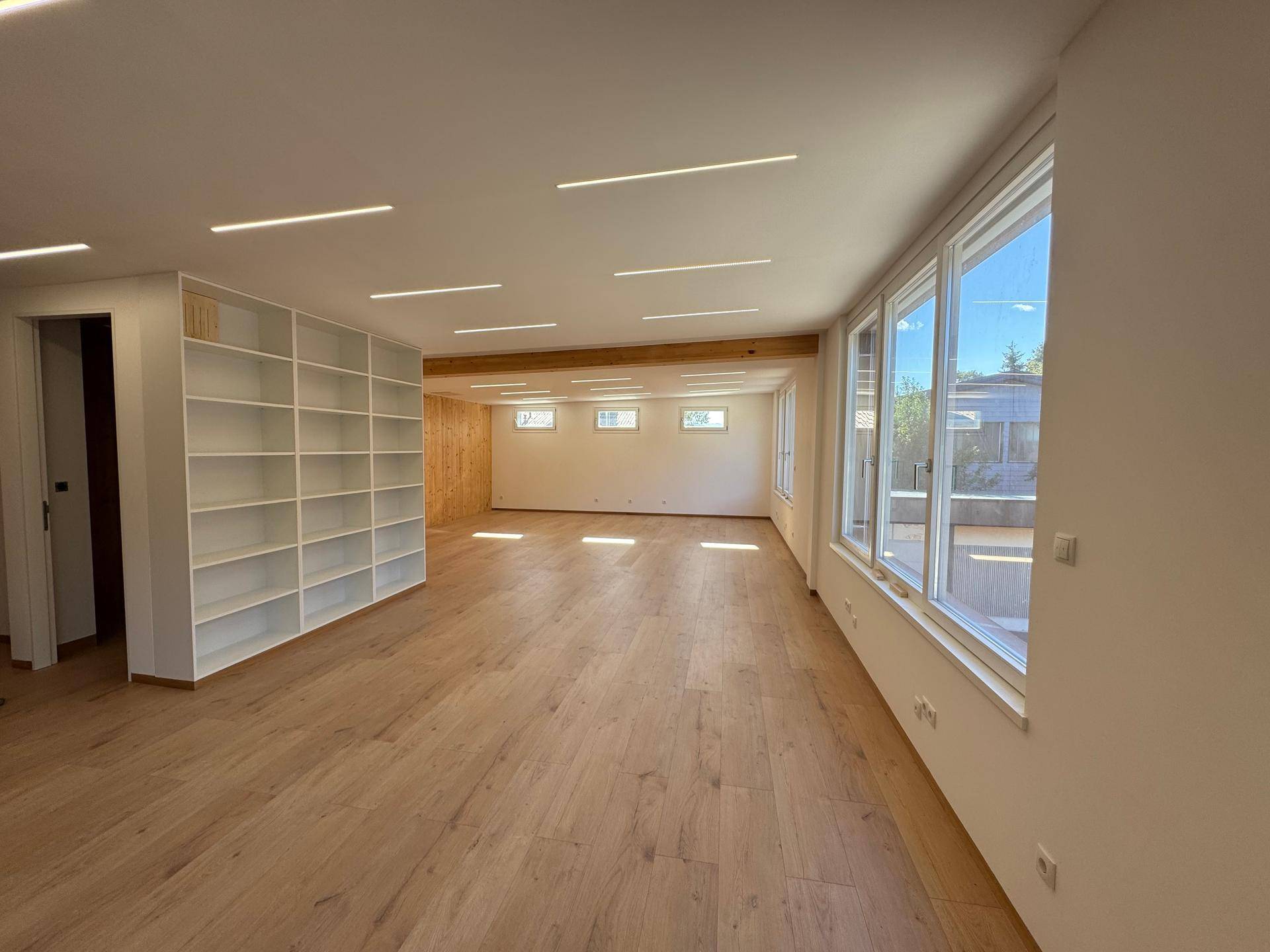 Großer, heller Büroraum mit Holzboden, langen Fensterfronten und integrierten Deckenleuchten.
