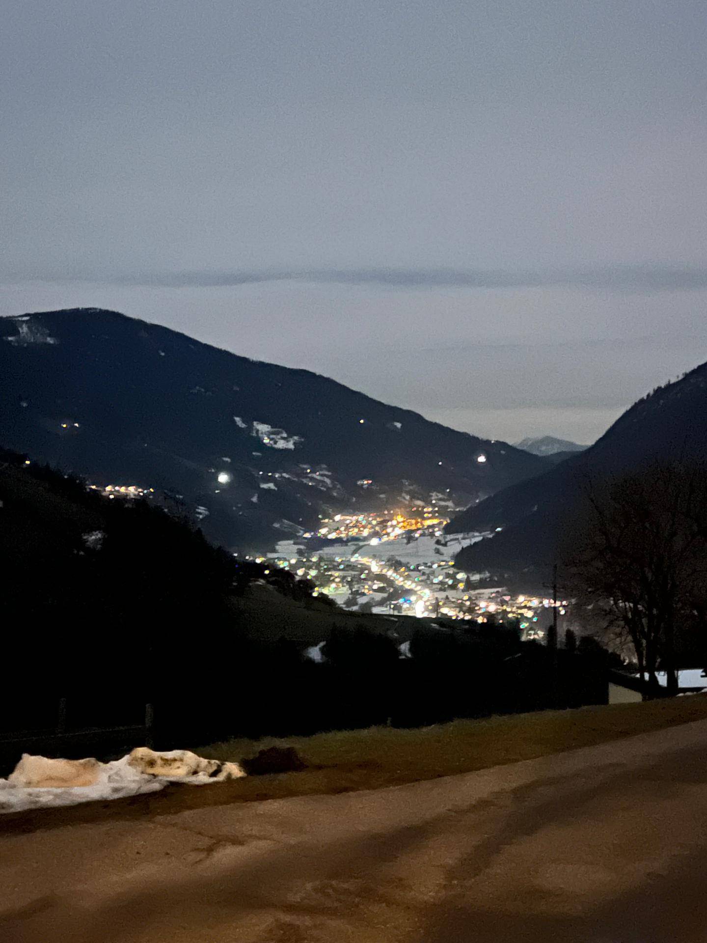 Nachtaufnahme eines beleuchteten Dorfes in einem verschneiten Tal, umgeben von dunklen Bergen.
