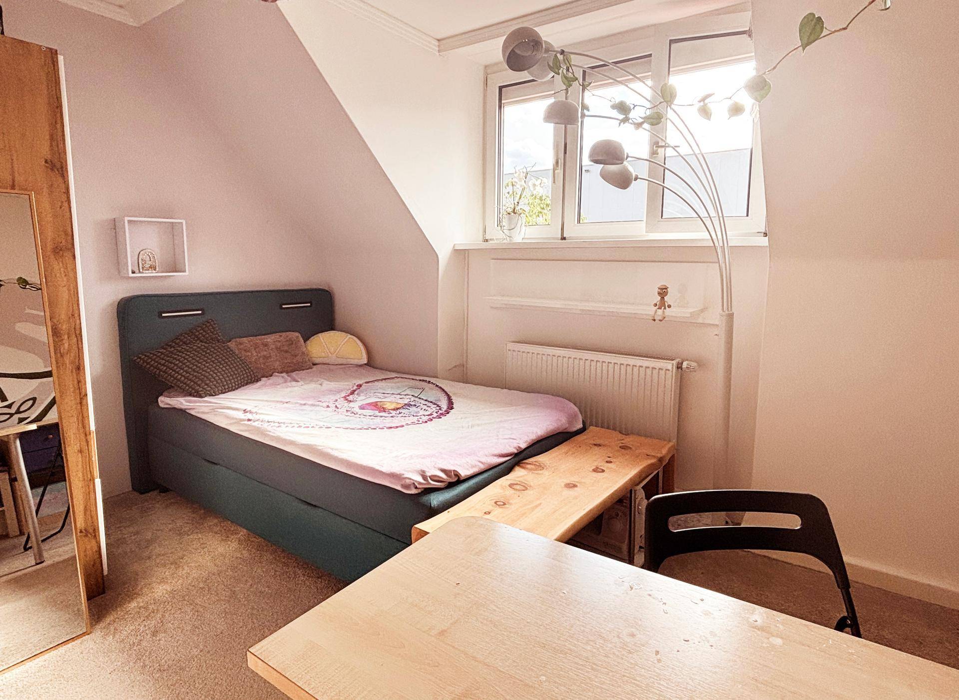 Helles Schlafzimmer mit Bett, Schreibtisch und Dachschräge, gut beleuchtet durch ein Fenster.