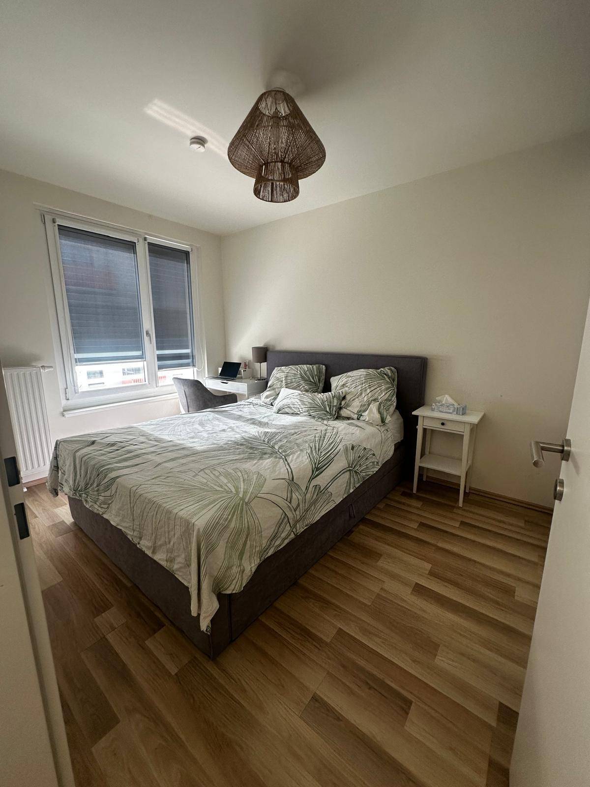 Helles Schlafzimmer mit Bett, Schreibtisch und Fenster mit Jalousien.