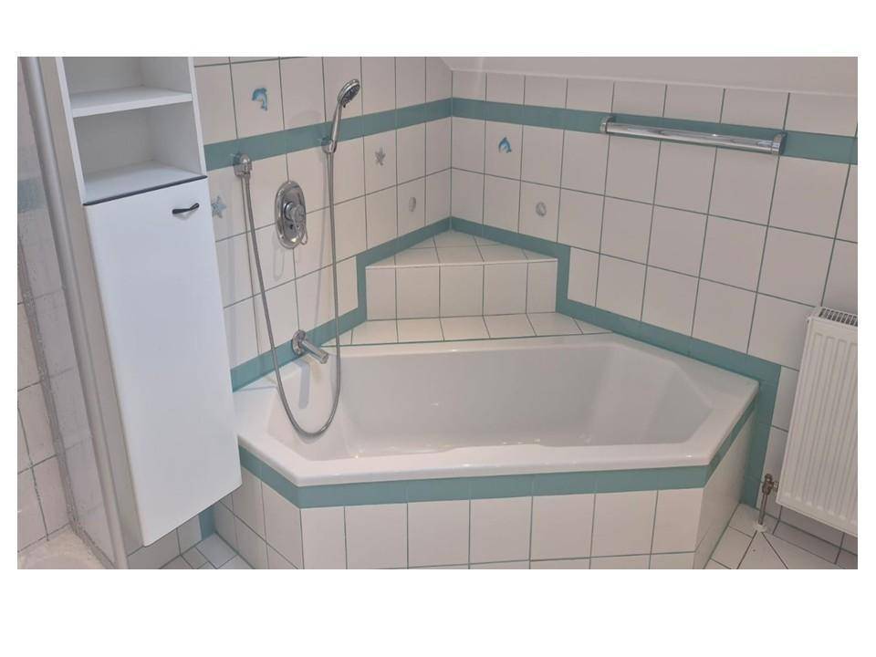 Eckbadewanne im gefliesten Badezimmer mit integrierter Duscharmatur und blauer Akzentlinie.