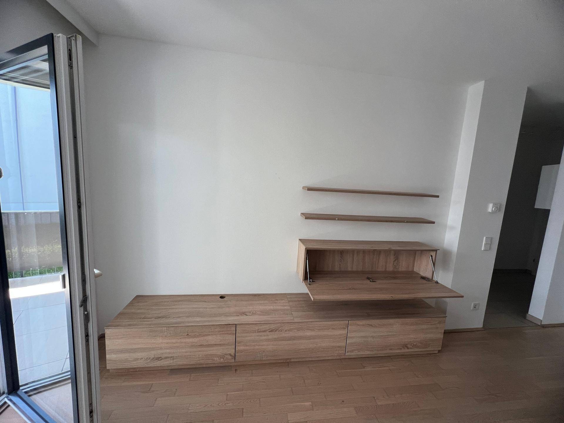 Wohnbereich mit modernen Holzmöbeln und Regalen, heller Raum mit Balkonzugang.
