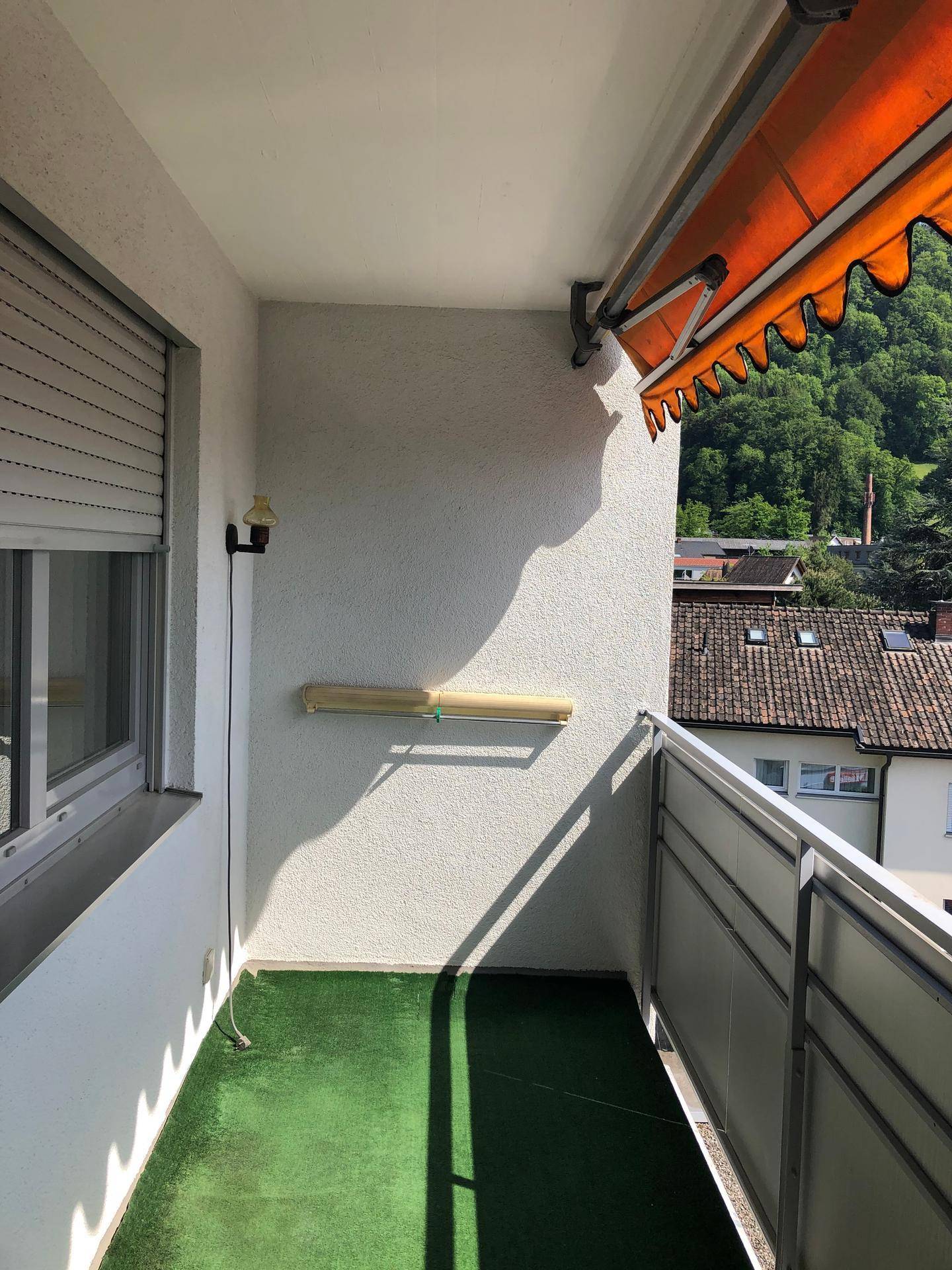 Sonniger Balkon mit Kunstrasen, Markise und Blick auf die Dächer der umliegenden Häuser.