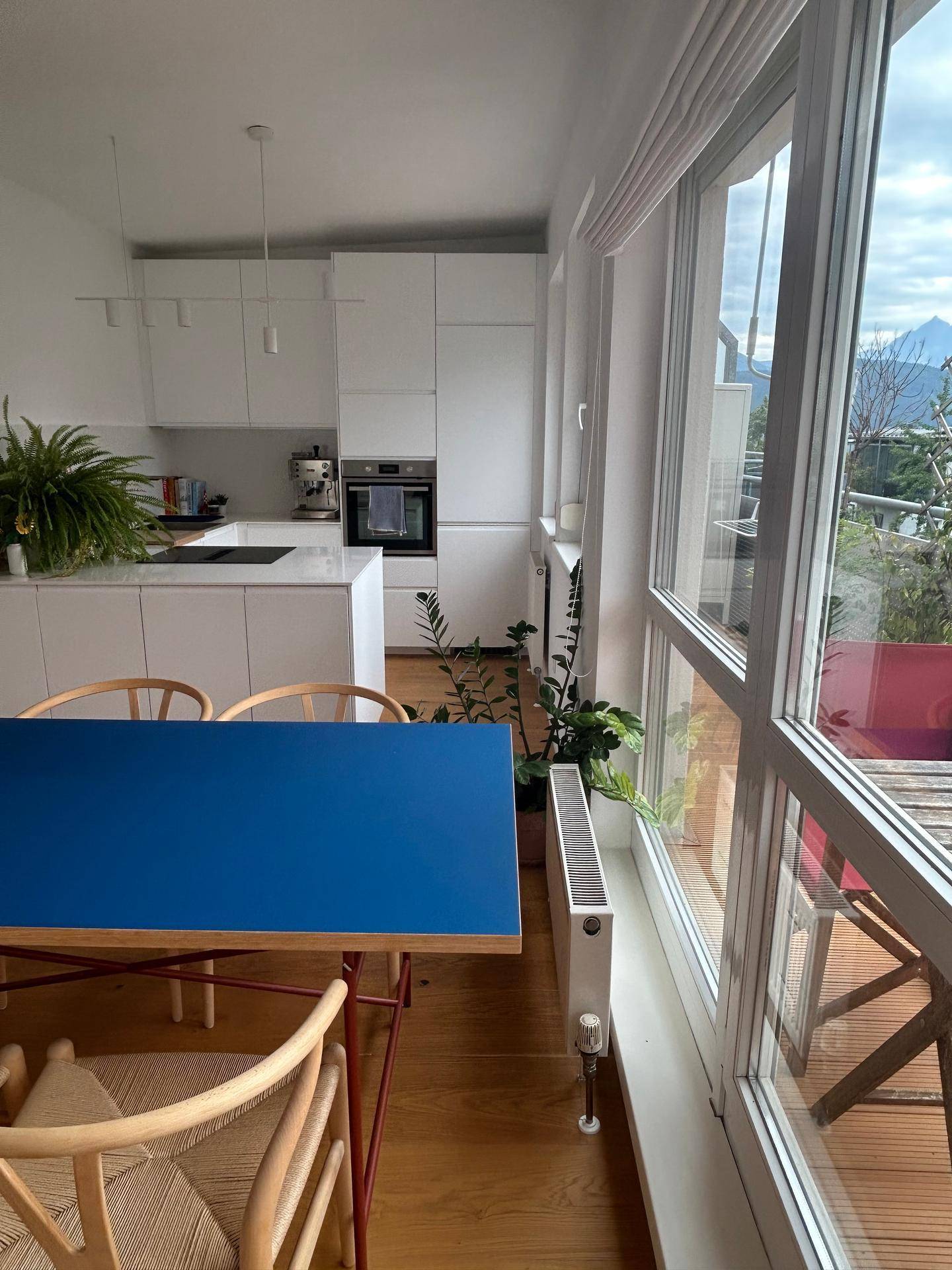 Moderner Essbereich mit blauem Tisch und offener, weißer Küche, umgeben von großen Fenstern.
