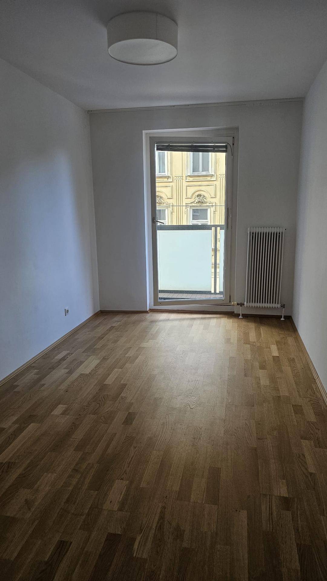 Leeres Zimmer mit Parkettboden und Zugang zu einem französischen Balkon mit Stadtblick.