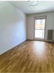 Helles Zimmer mit Parkettboden, weißen Wänden und Zugang zu einem kleinen Balkon.