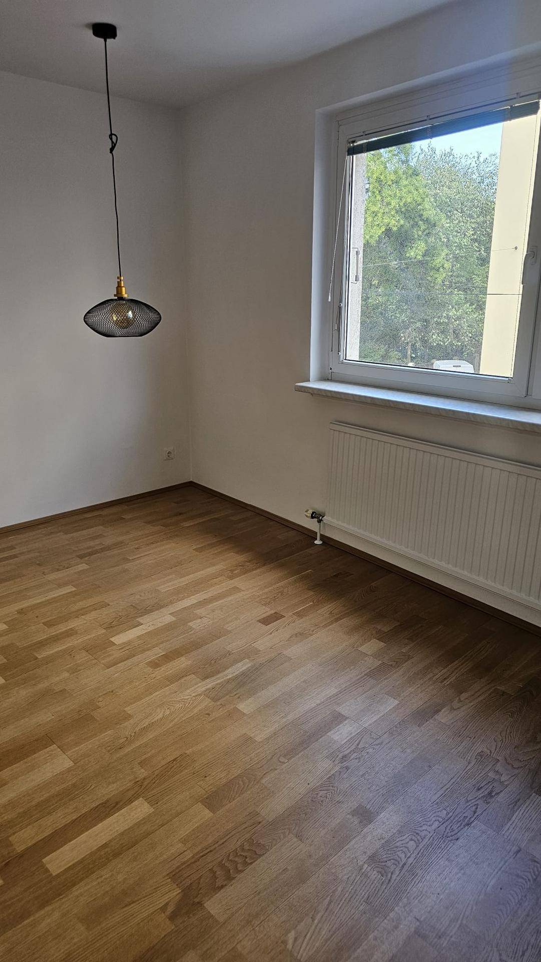 Helles, leeres Zimmer mit Parkettboden, Fensterblick ins Grüne und einer modernen Hängelampe.