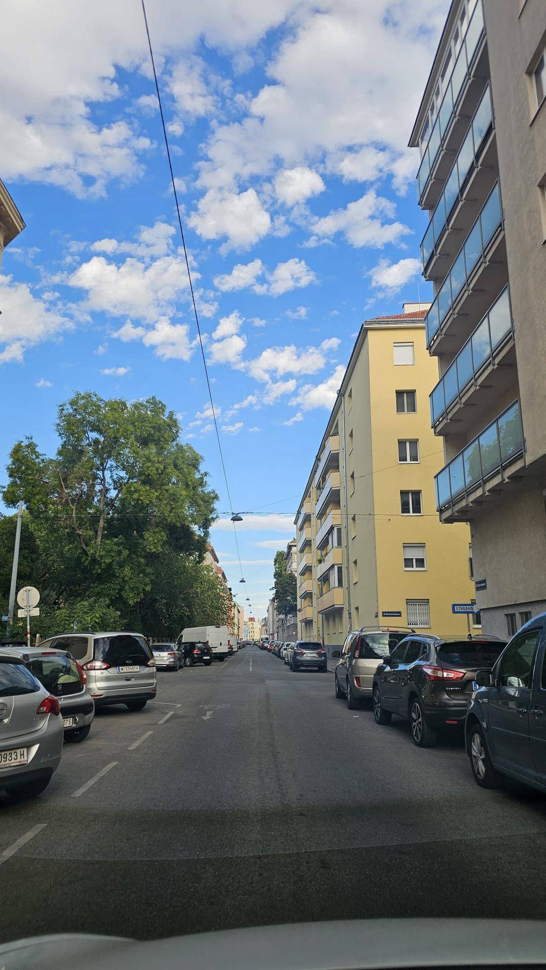 Eine belebte Straße mit parkenden Autos, umgeben von Gebäuden und Bäumen unter blauem Himmel.