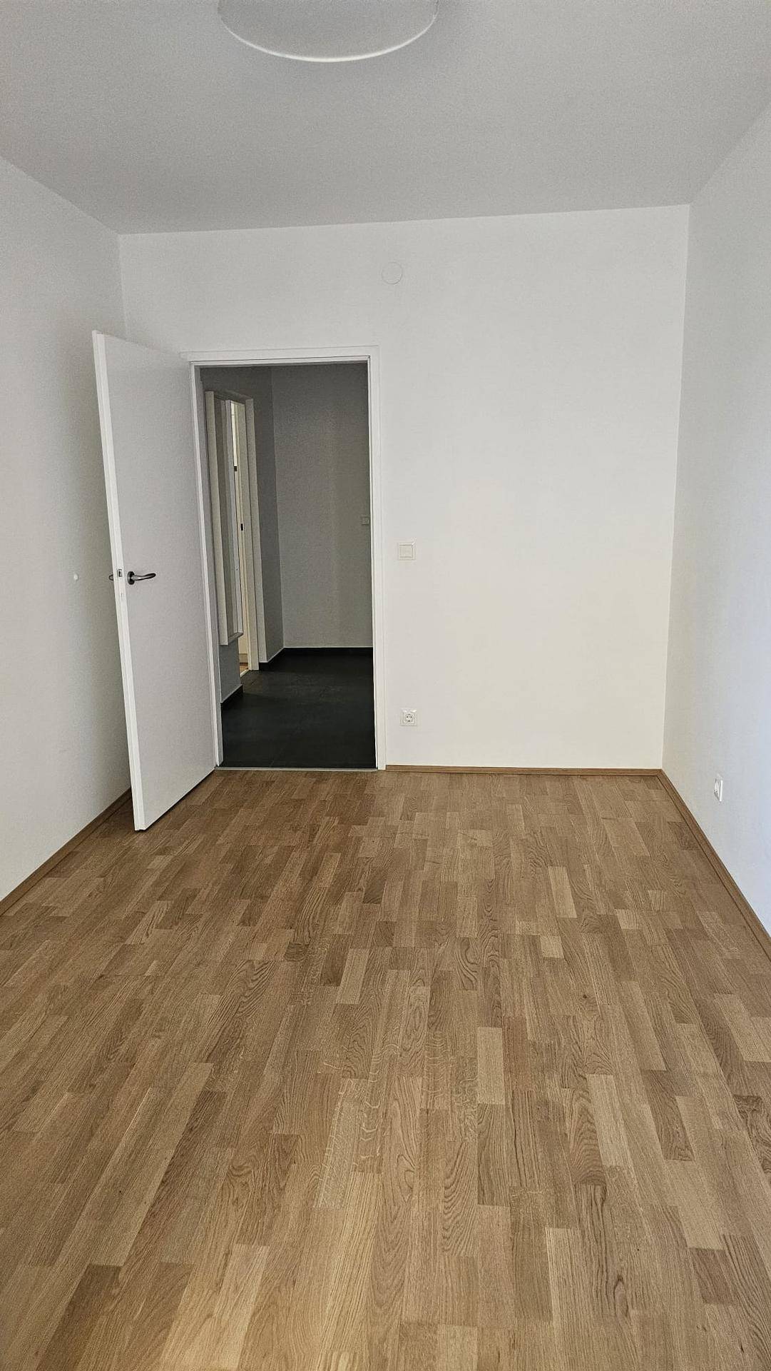 Helles, leeres Zimmer mit Parkettboden und Zugang zu einem Flur mit dunklen Fliesen.