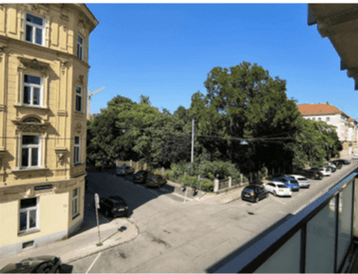 Weitblick vom Balkon auf eine belebte Straßenecke mit Bäumen und Stadtgebäuden.