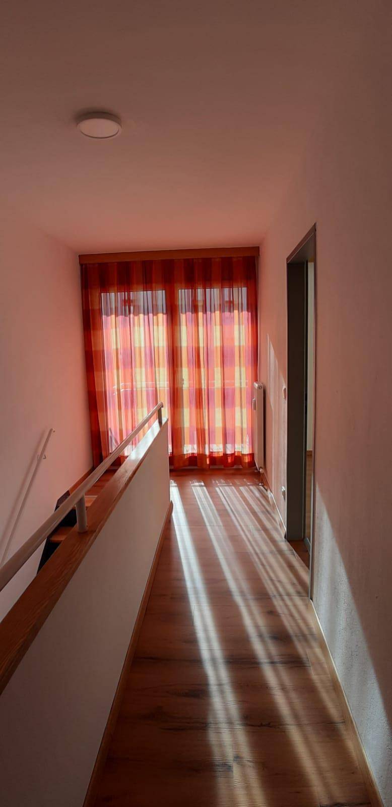 Lichtdurchfluteter Flur im Obergeschoss mit Holzboden und Fenster mit bunten Vorhängen.