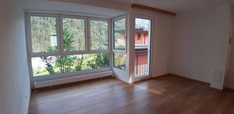Heller Wohnraum mit großen Fenstern, Holzboden und Blick auf die grüne Umgebung.