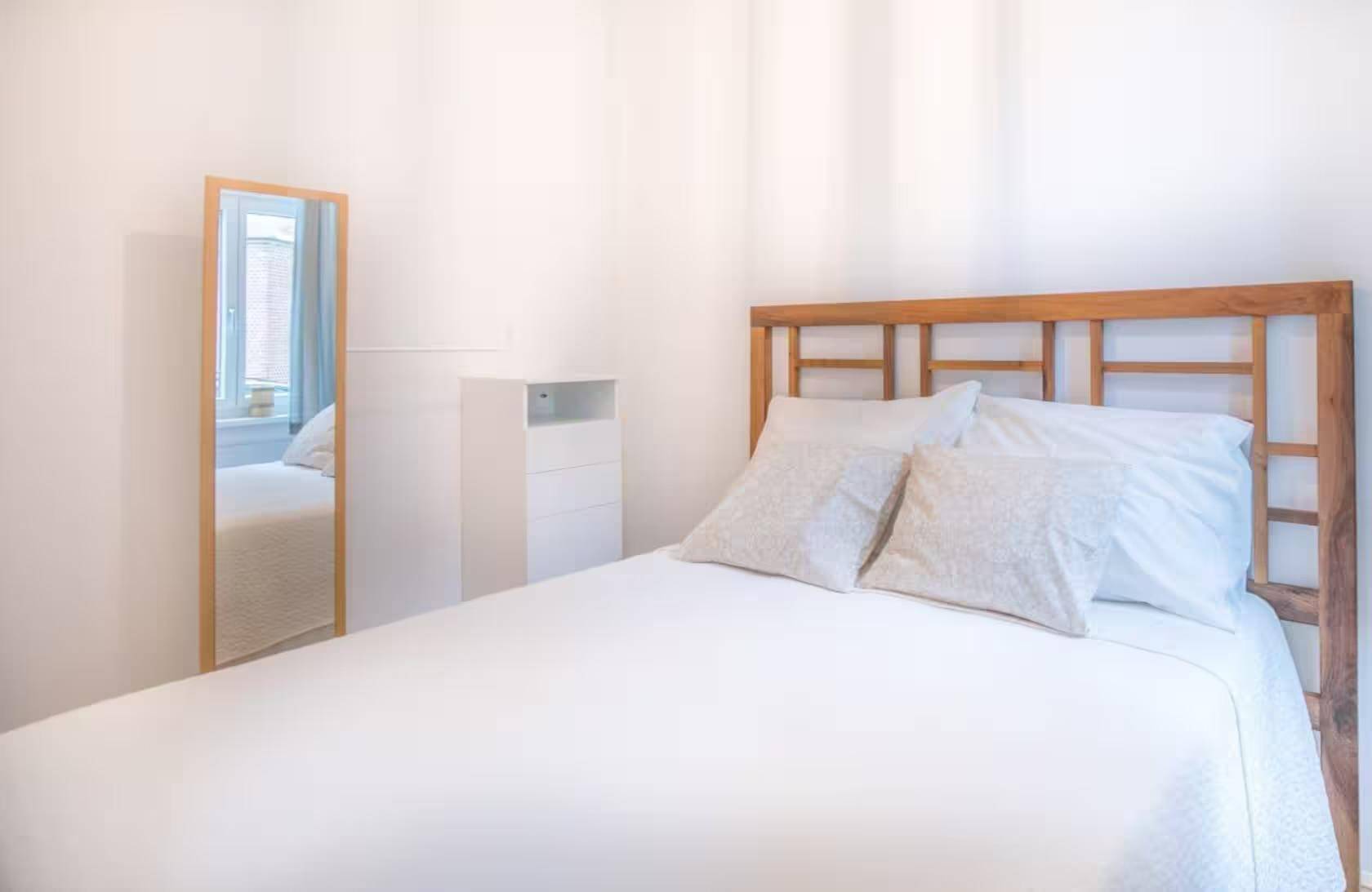 Helles Schlafzimmer mit Holzbettrahmen, weißer Bettwäsche und großem Standspiegel.