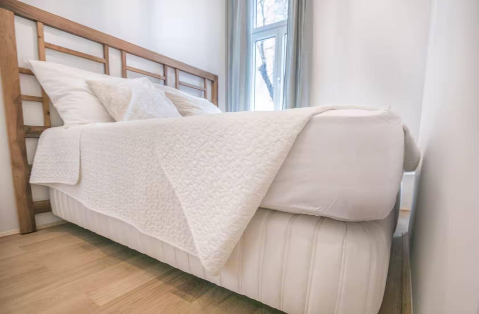 Gemütliches Bett mit weißer Bettwäsche und Holzkopfteil auf hellem Holzboden.